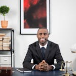 African American businessman smiling in an office environment, seated at a desk.